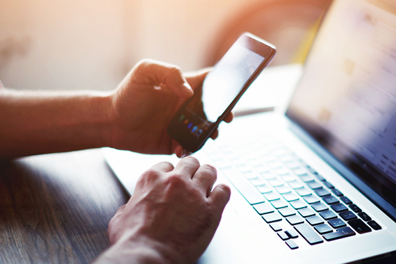 Ansicht von zwei Händen einer Person auf einem Tisch. Die eine Hand hält ein Handy, die andere bewegt den Cursor auf einem Laptop. 
